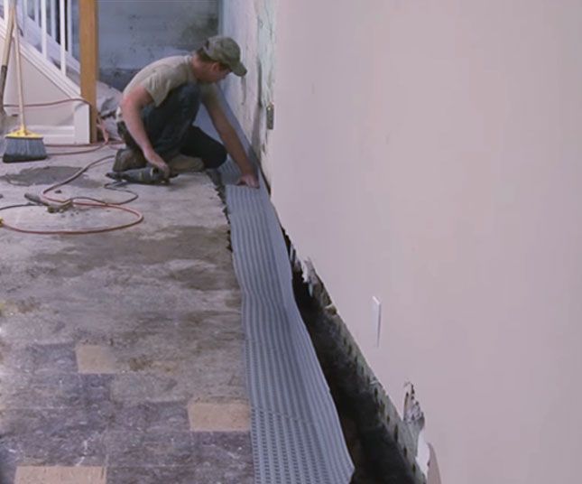 Worker installing a long gray drainage channel along a wall on a concrete floor.