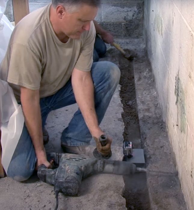 Worker using a power jackhammer to break concrete in a narrow trench beside a wall