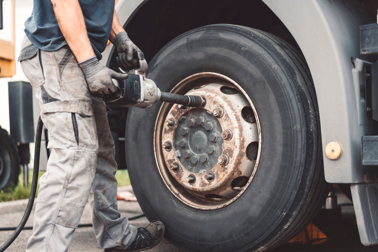 Mechanic kneeling, removing lug nuts from a truck tire with a wrench. Outdoors, blue work clothes.