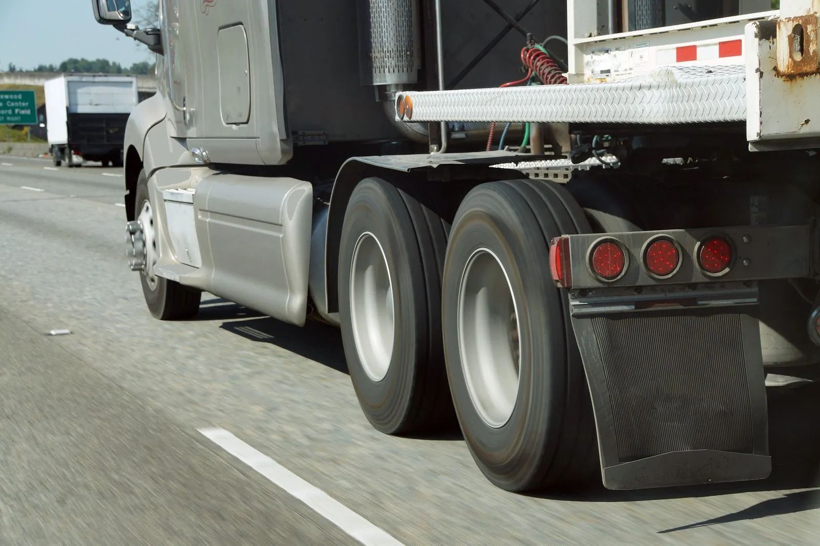 Close-up of a semi-truck's rear wheels and trailer, driving on a highway, with red tail lights visible.