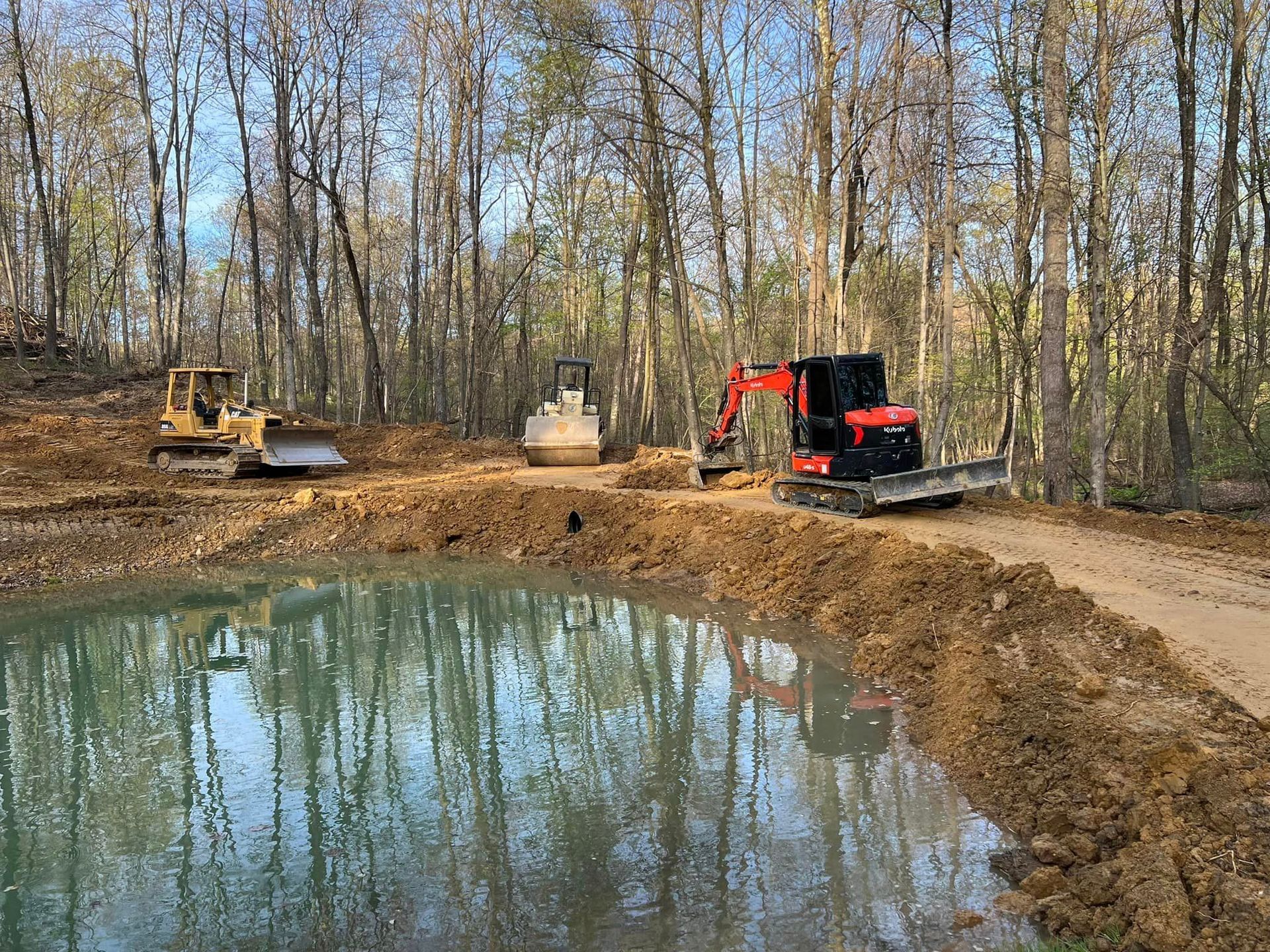 A couple of tractors are working on a pond in the woods.