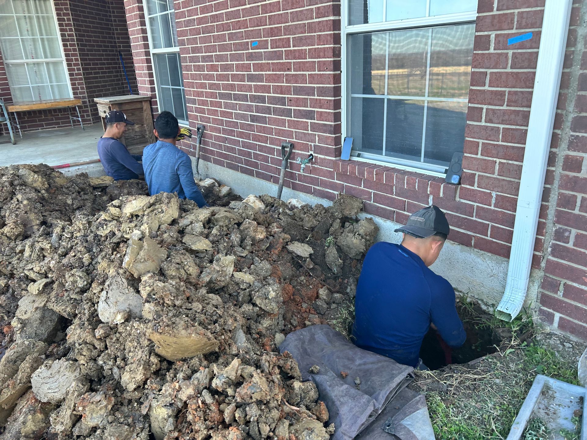 Two men are digging a hole in the ground in front of a brick house.
