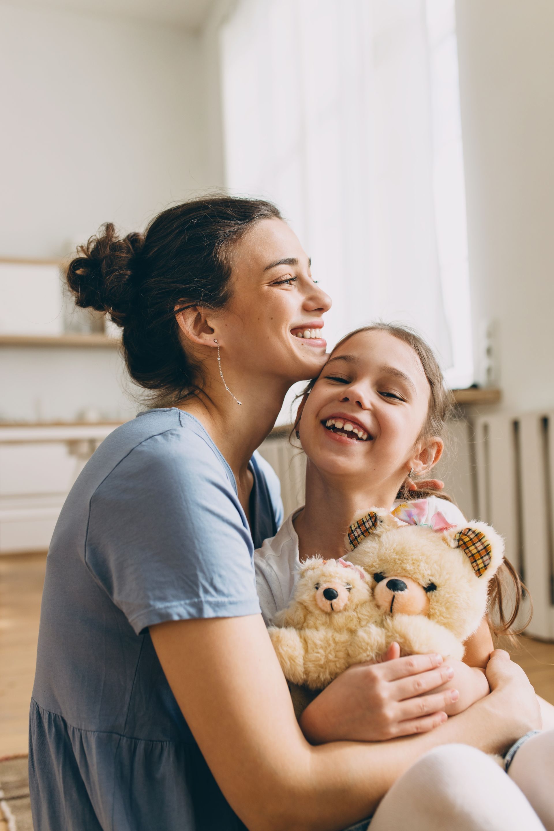 A woman is kissing a little girl on the cheek while holding a teddy bear.