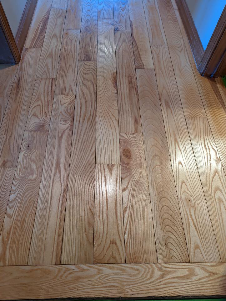 Wooden floorboards, light-colored wood, in a hallway. Boards are laid in a staggered pattern.