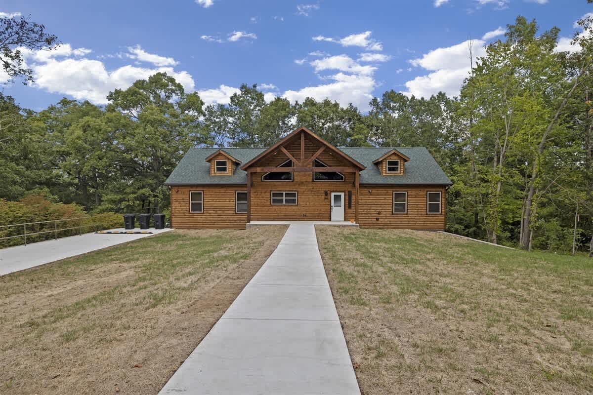A large log cabin with a concrete driveway leading to it is surrounded by trees.