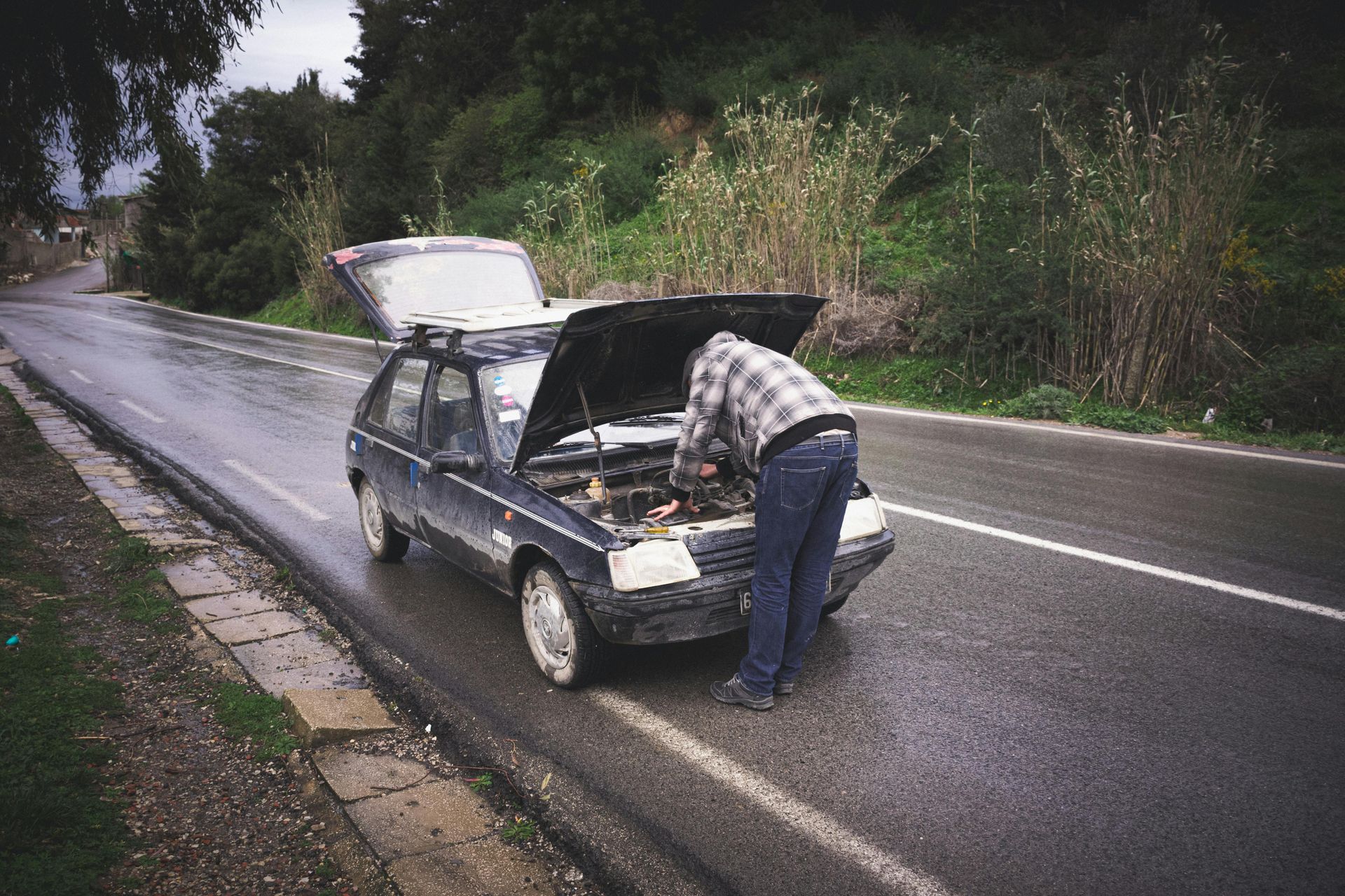 A man is looking under the hood of a broken down car.