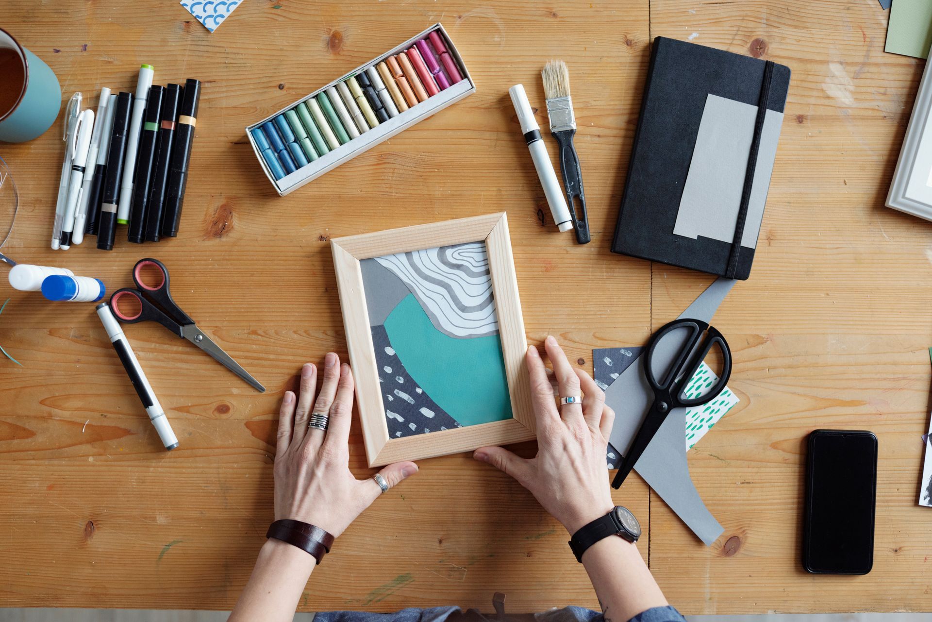 Hands arranging a framed art print on a desk with paints, scissors, notebook, and phone.