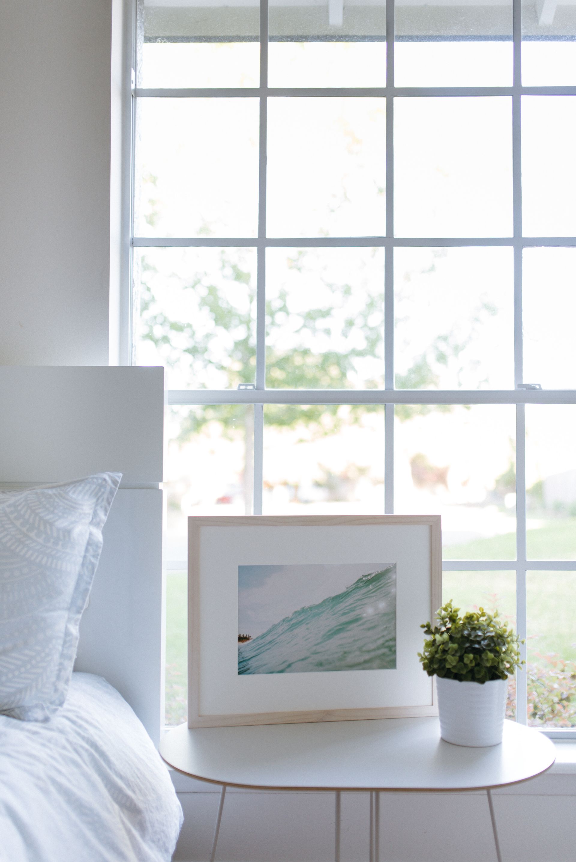 Cozy bedroom corner with a bed, round white table, framed art, and potted plant by a bright window