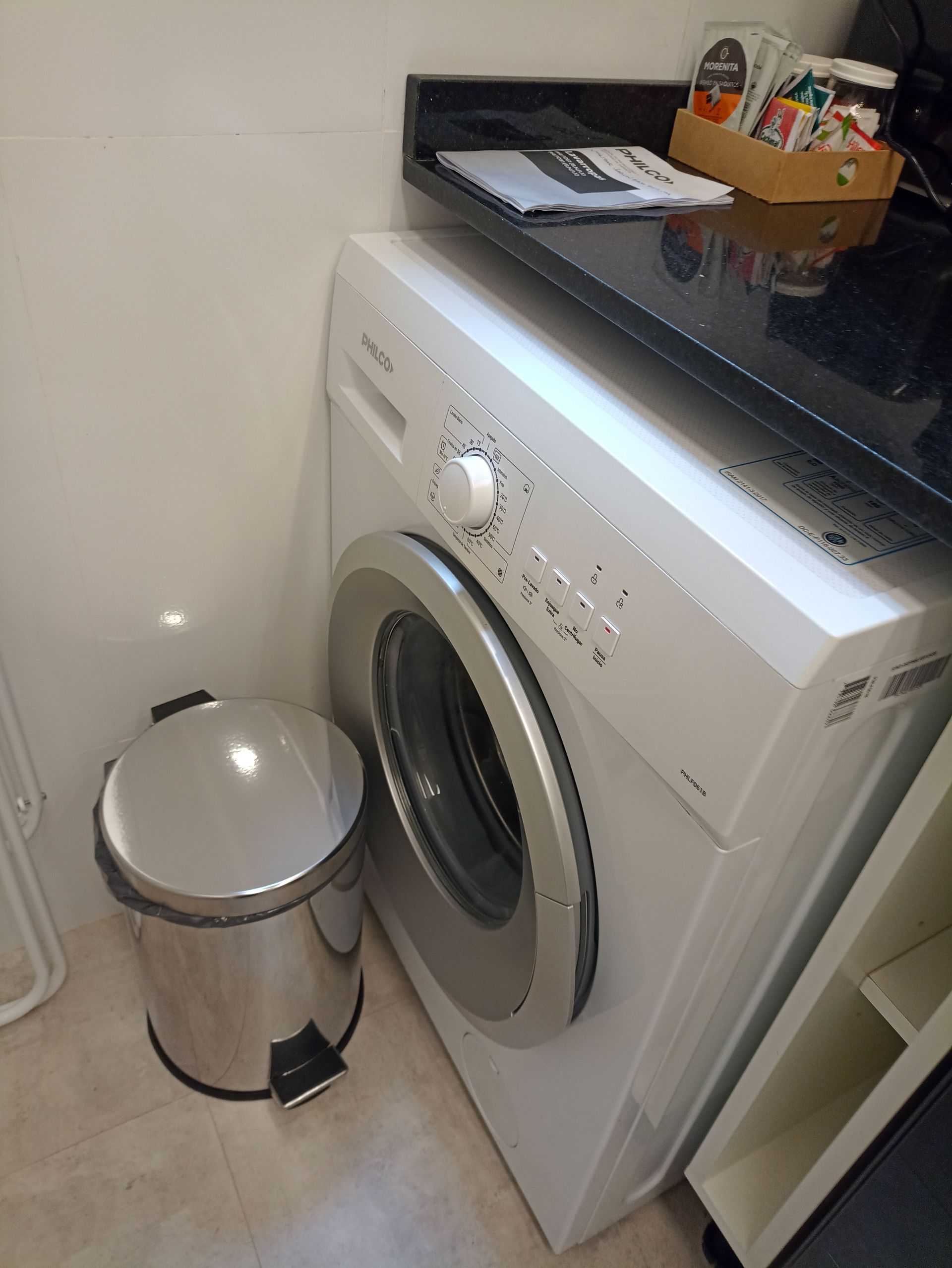 Una lavadora está al lado de un bote de basura en un lavadero.