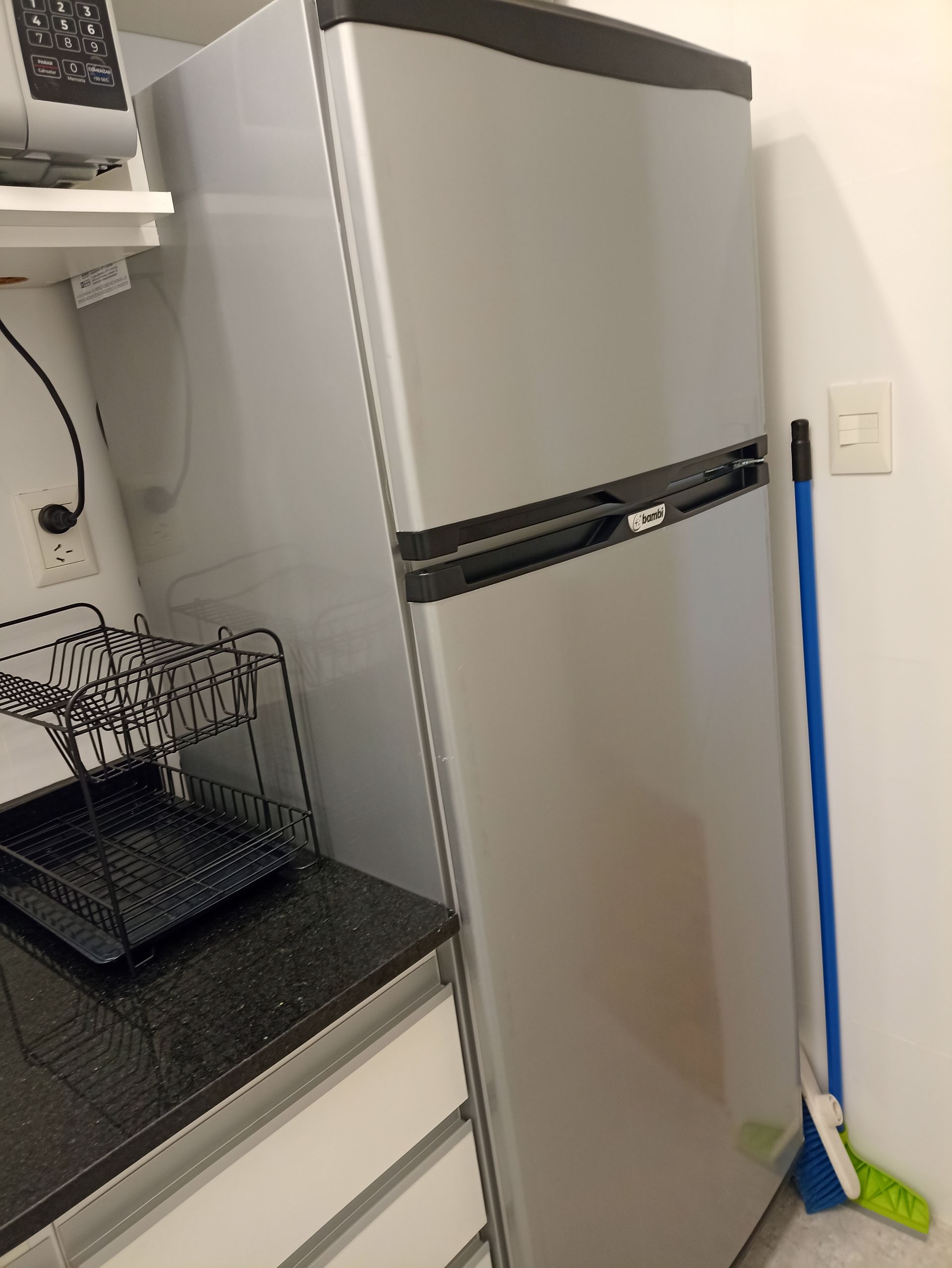 En una cocina hay un refrigerador de acero inoxidable al lado de una escoba.