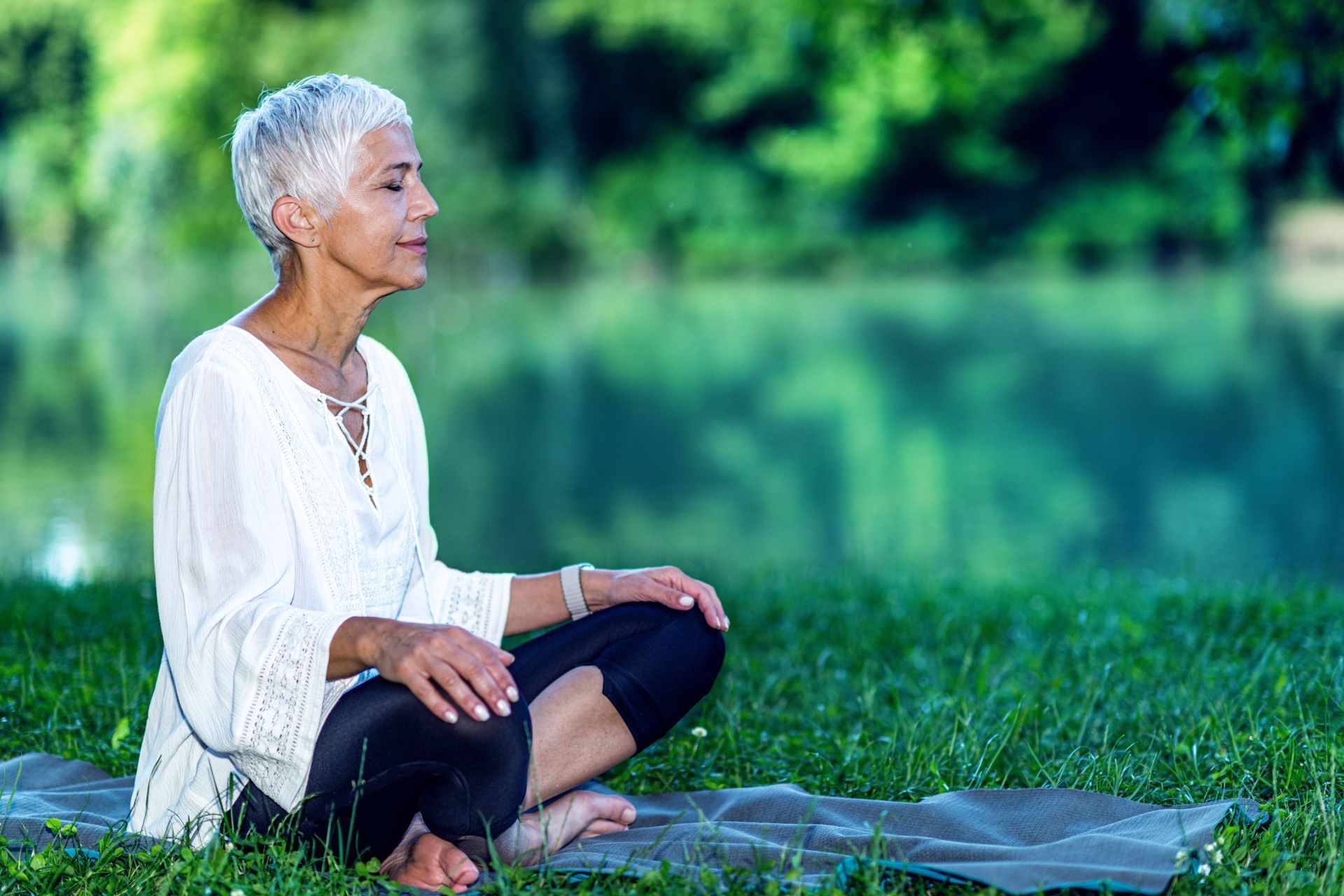 An older woman is sitting on a yoga mat in the grass.