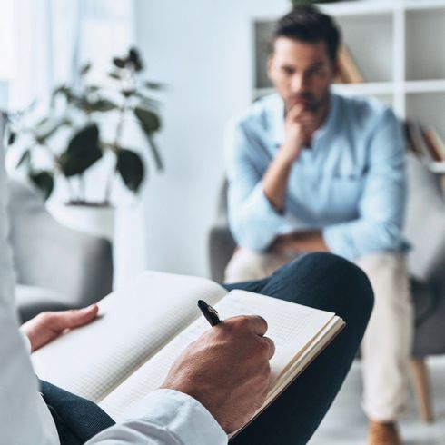 A man is sitting in a chair while a person writes in a notebook.