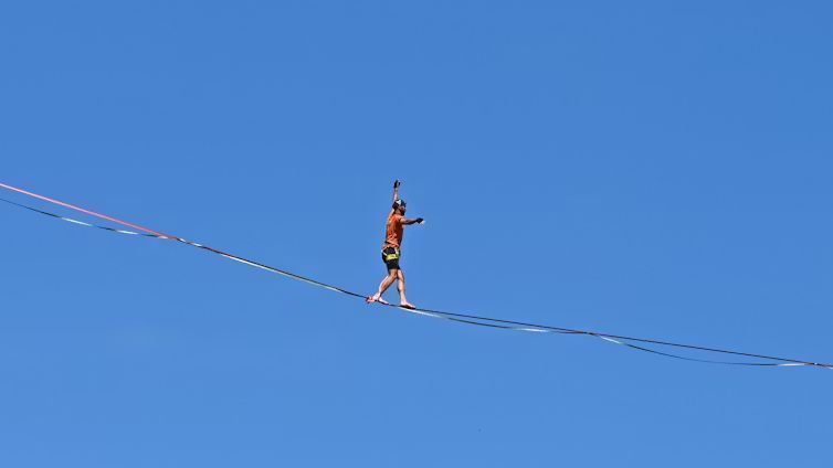 A man wearing an orange shirt and safety gear balances carefully while walking across a high slackli