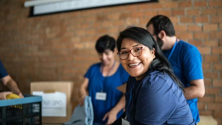 A smiling woman wearing glasses and a volunteer lanyard looks toward the camera while other individuals sort donation boxes.