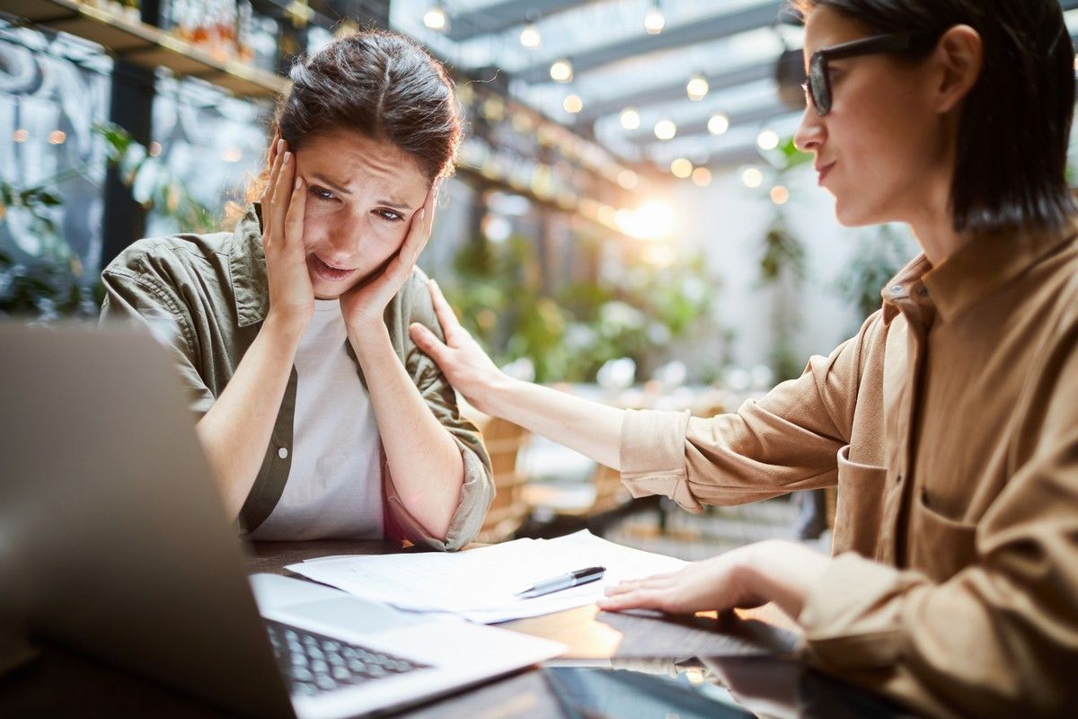 A woman is comforting another woman who is sitting at a table with a laptop.