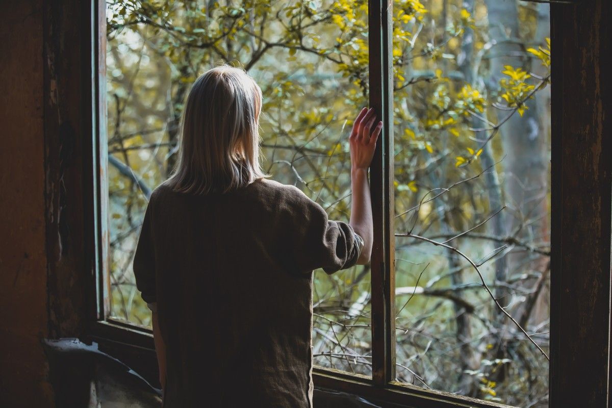 Woman gazing out a window into a wooded area, hand resting on the glass.