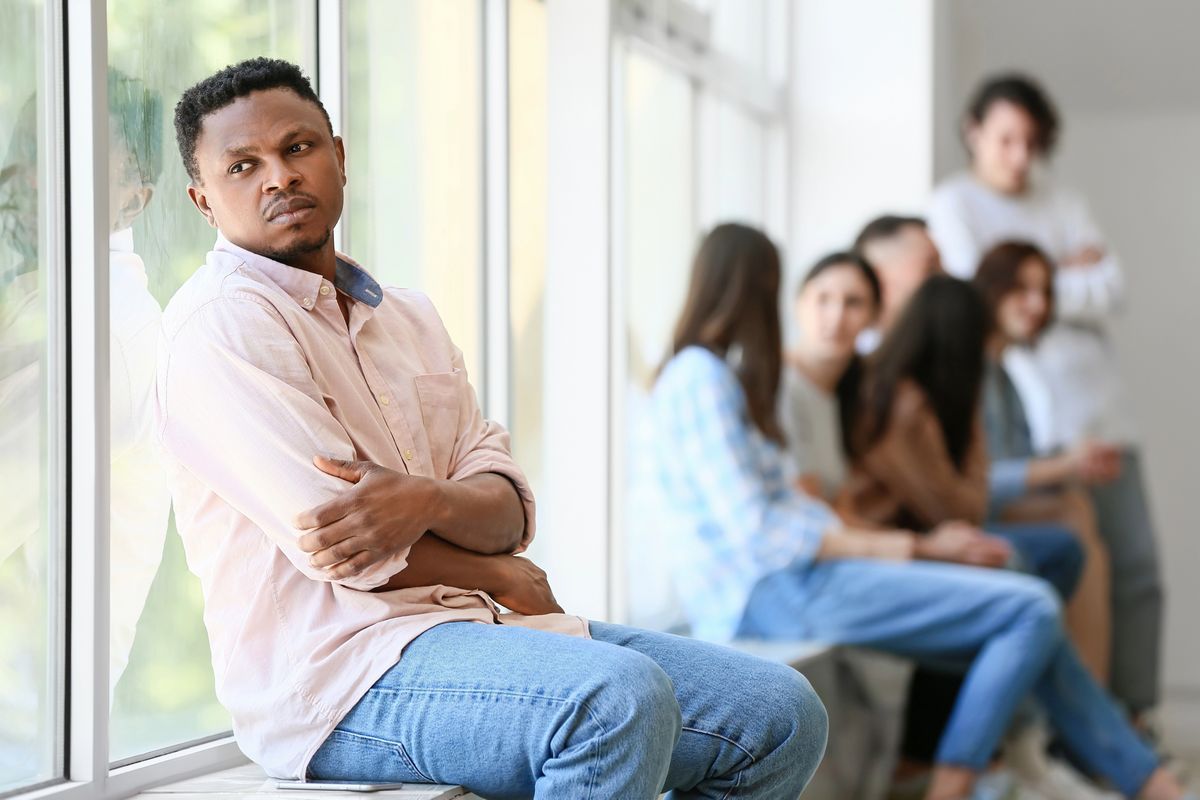 Man sitting apart from group, looking upset, representing effects of discrimination on mental health.