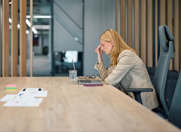 A blonde woman wearing a beige blazer sits at a long wooden office desk with a laptop, pinching the