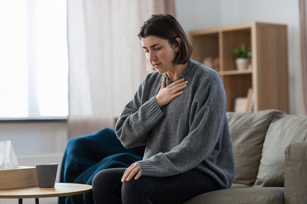 Woman sitting on a couch with hand on chest, appearing anxious or overwhelmed.