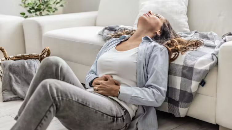 A woman sitting on the floor clutching her stomach with an expression of severe physical and emotional distress from PMDD.