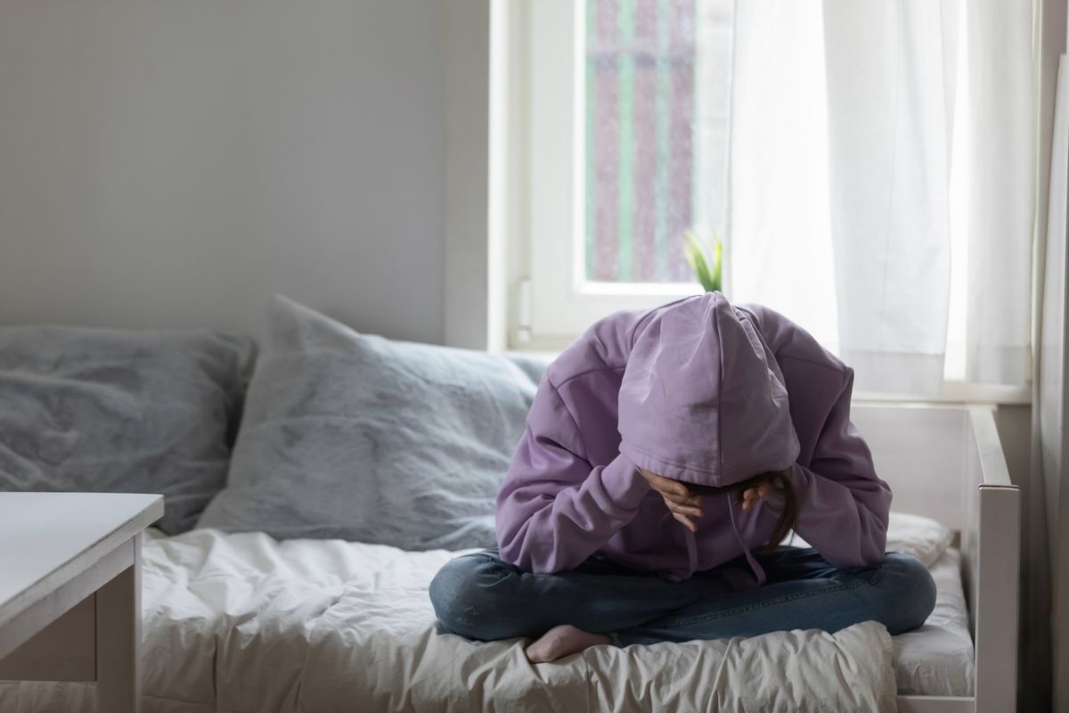 Teen sitting on bed with head in hands, symbolizing trauma’s impact on brain development and emotional coping in teens.