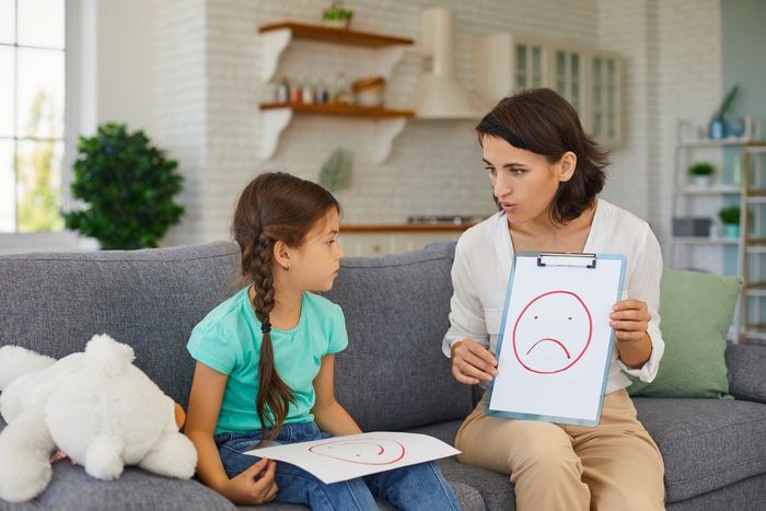 A woman is sitting on a couch with a little girl and holding a clipboard with a sad face drawn on it.
