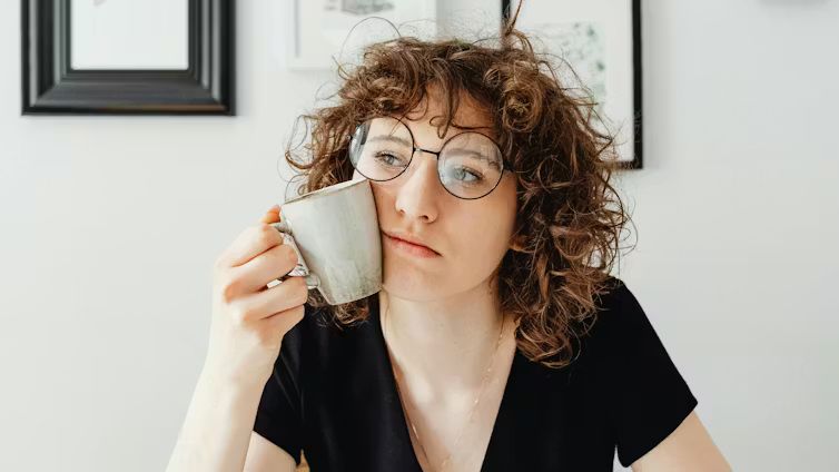 A woman with curly hair and glasses holding a coffee mug to her cheek with a deeply pensive, analytical expression.