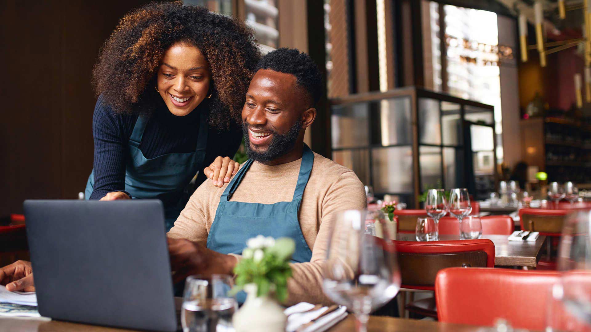 Restaurantbesitzer schauen lächelnd gemeinsam auf einen Laptop. Eine Frau beugt sich über die Schulter eines Mannes. Im Hintergrund sind Tische gedeckt.