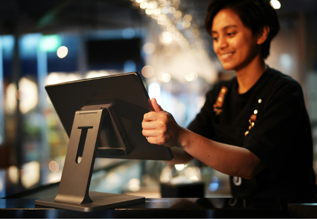 Eine Person lächelt bei der Bedienung eines Touchscreen-Kassensystems. Schwarzer Kassenständer vor dunklem Hintergrund.