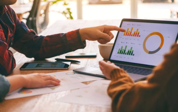 Two people reviewing charts and graphs on a laptop and smartphone at a table.
