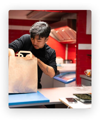 Ein Koch in schwarzem Hemd packt an einer Theke mit rot-weißer Innenausstattung eine Tüte zum Mitnehmen.