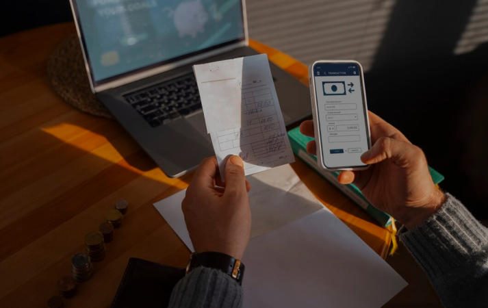 Hände halten eine Quittung und ein Telefon. Laptop und Papier liegen auf dem Tisch in einem hell erleuchteten Raum.