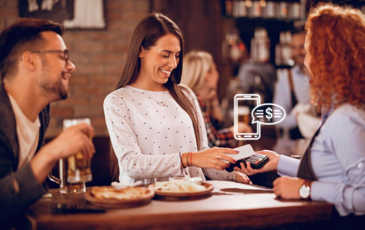 Frau bezahlt an einer Bar mit dem Handy, lächelt, Mann neben ihr trinkt Bier, Kellner hält ein Zahlungsgerät.