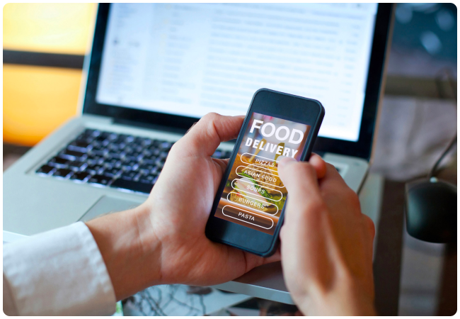 Eine Person nutzt eine Essensliefer-App auf ihrem Smartphone; im Hintergrund ist ein Laptop zu sehen.