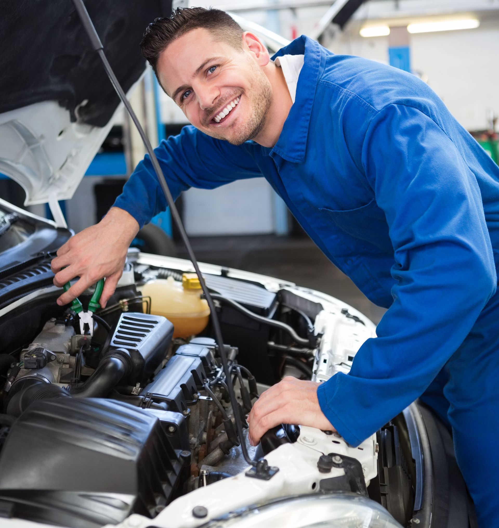 A smiling mechanic is working under the hood of a car in a garage.