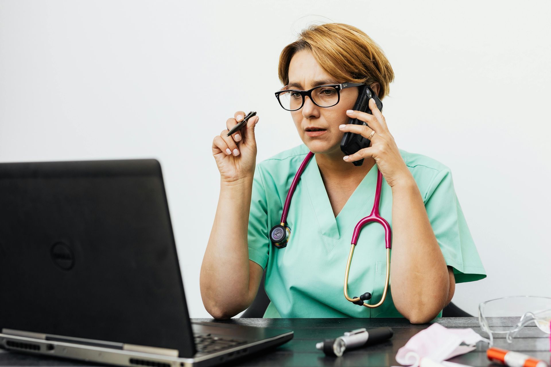 Healthcare worker in scrubs on phone, looking at laptop, stethoscope, pen in hand.
