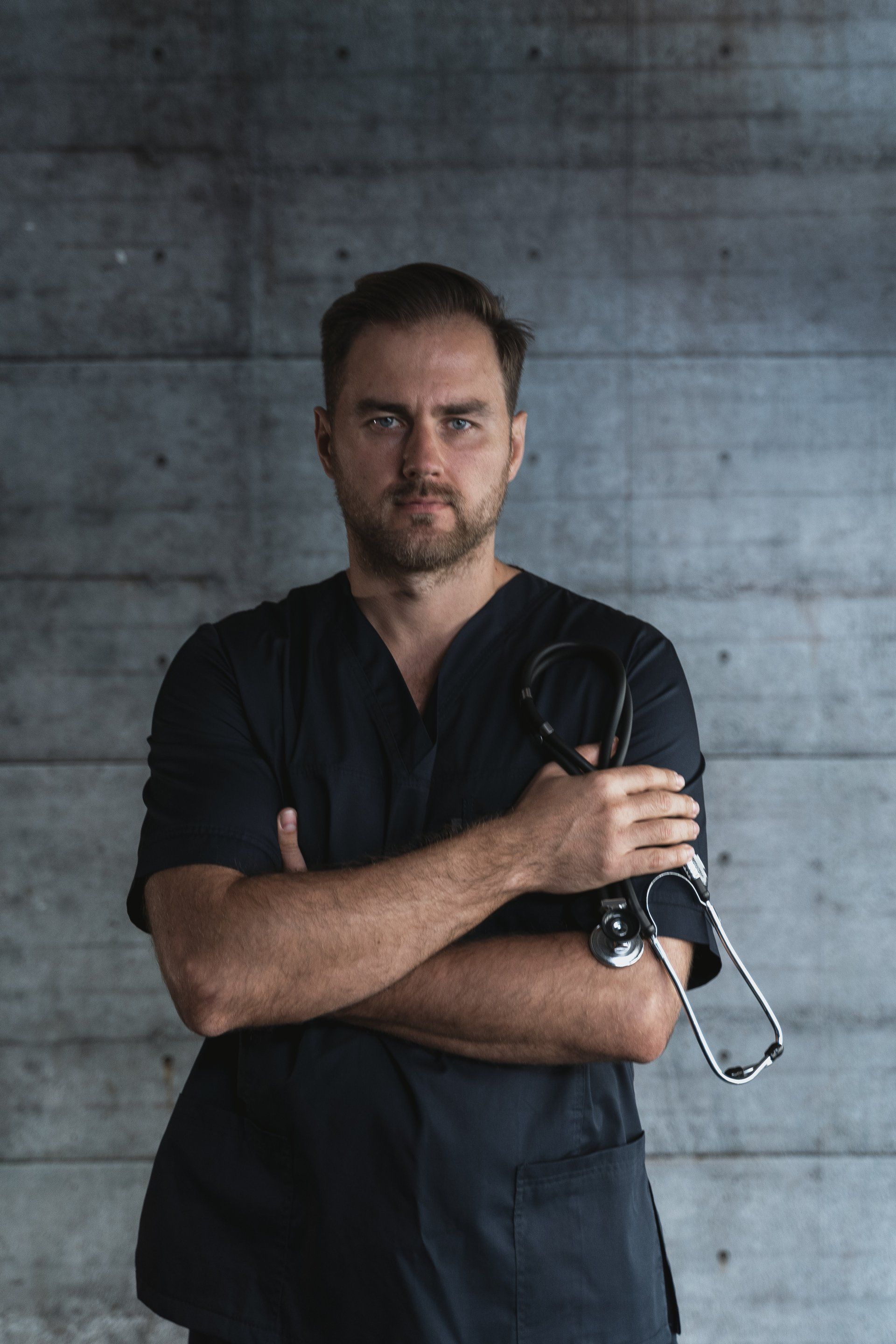 Man in black scrubs, arms crossed, holding stethoscope; gray textured wall background.