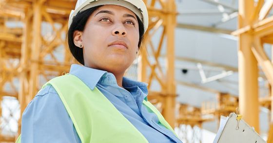 Eine Frau mit Schutzhelm und Sicherheitsweste steht mit einem Klemmbrett in der Hand vor einer Baustelle.