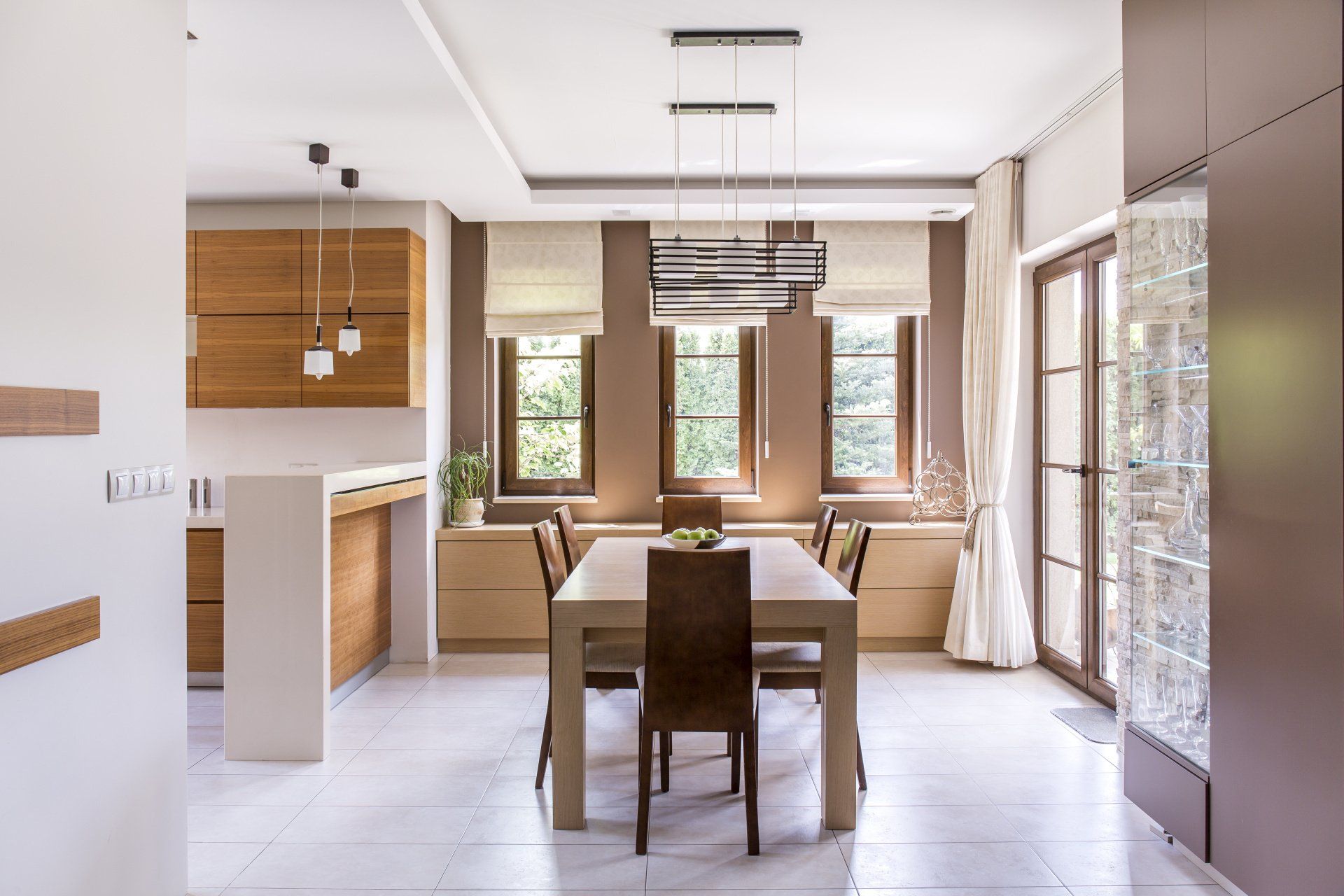 a dining room with a table and chairs and a kitchen in the background and windows with roman shades Simply Windows.