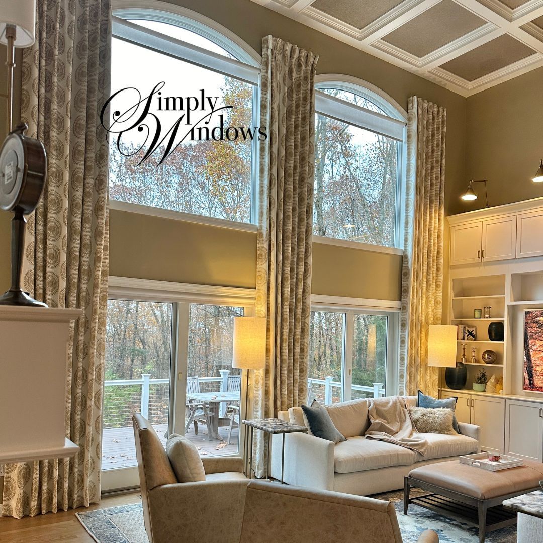 Bright living room with tall windows, patterned drapery, beige sofa, ottoman, and built-in shelves