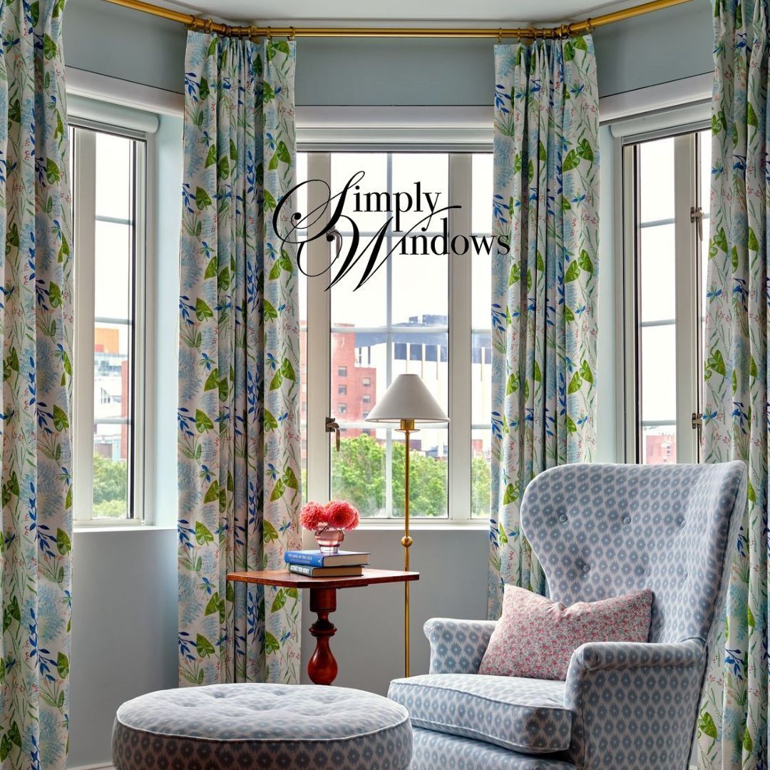A corner window nook with floral curtains, a wooden side table with a lamp, and a patterned blue armchair with ottoman.