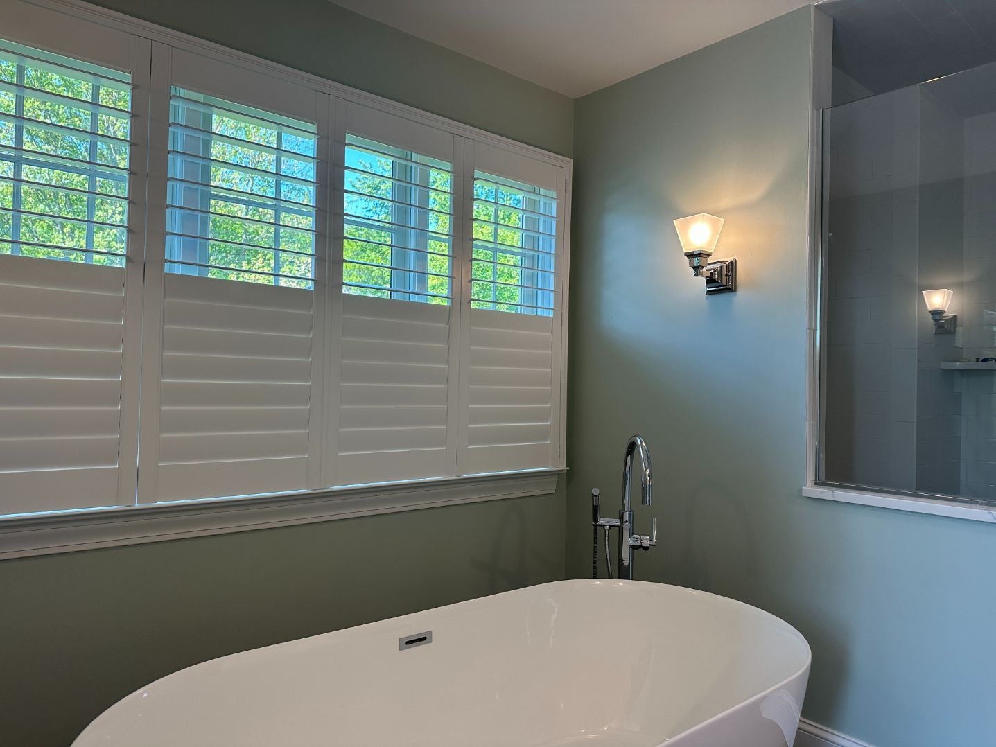 Bright bathroom with white shutters, wall sconces, and a freestanding tub by the window.