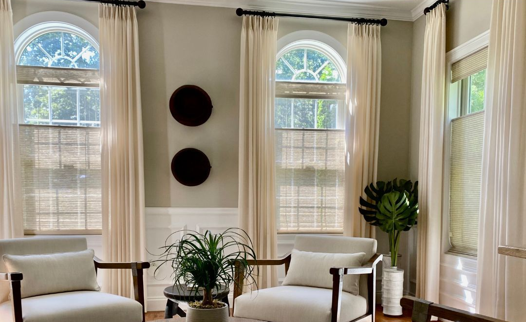Bright living room with tall windows, cream curtains, white chairs, and potted plants around a central wall art display