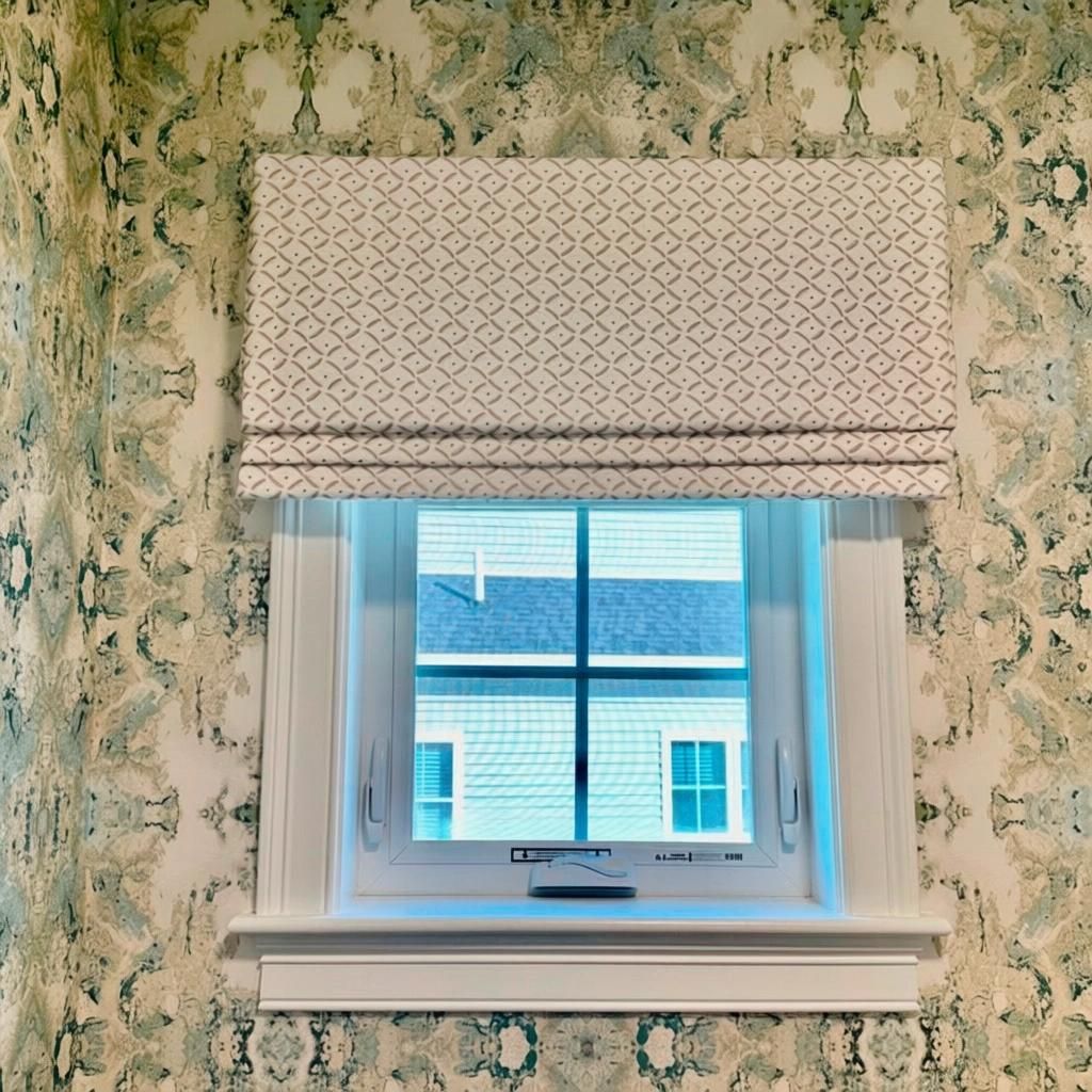 Window with white trim and a geometric print Roman shade, set against patterned wallpaper.