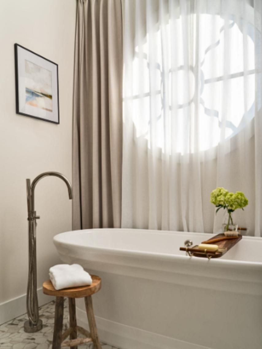 Bathroom with white tub, round window, gray curtains, wood stool, and framed art.
