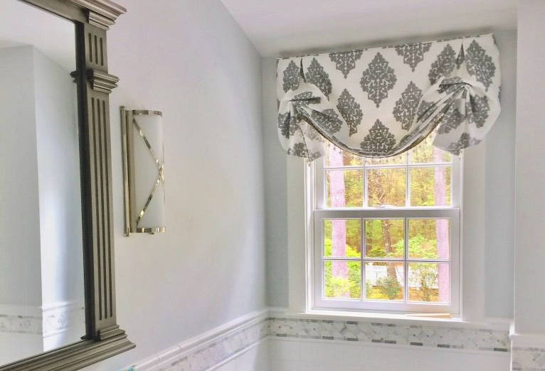 A bathroom vanity area with a decorative mirror, wall sconce, and a window covered by a patterned fabric valance.