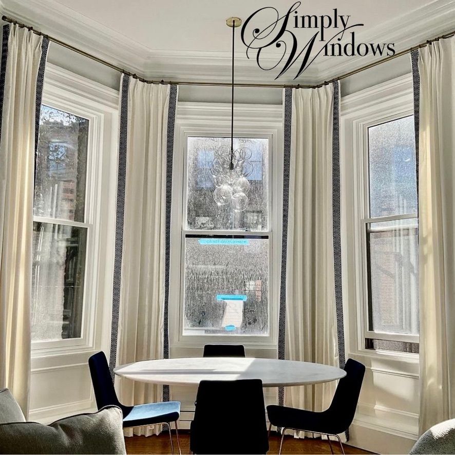 A dining area with a round white table, four black chairs, and a bay window framed by cream curtains with blue trim.