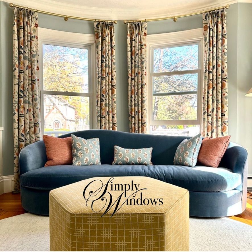 A curved navy sofa with patterned and solid pillows sits before a bay window with botanical drapes and a yellow ottoman.