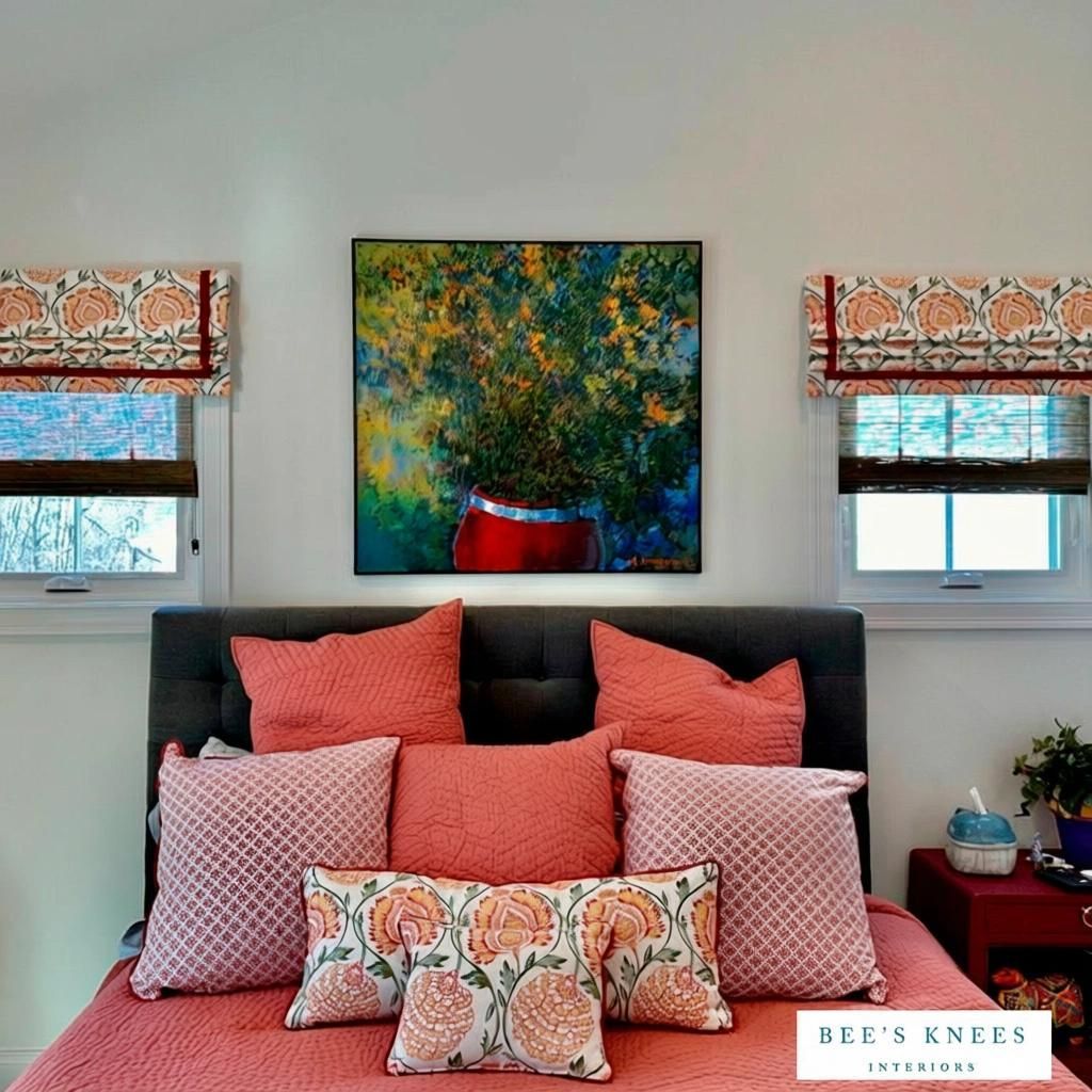 Bedroom with coral bedding, artwork, patterned Roman shades, and a red nightstand.