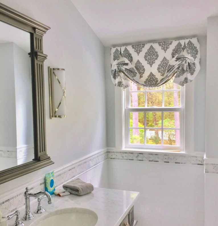 Bathroom with marble countertop and ornate window valance, featuring a mirror, sink, and window with greenery.